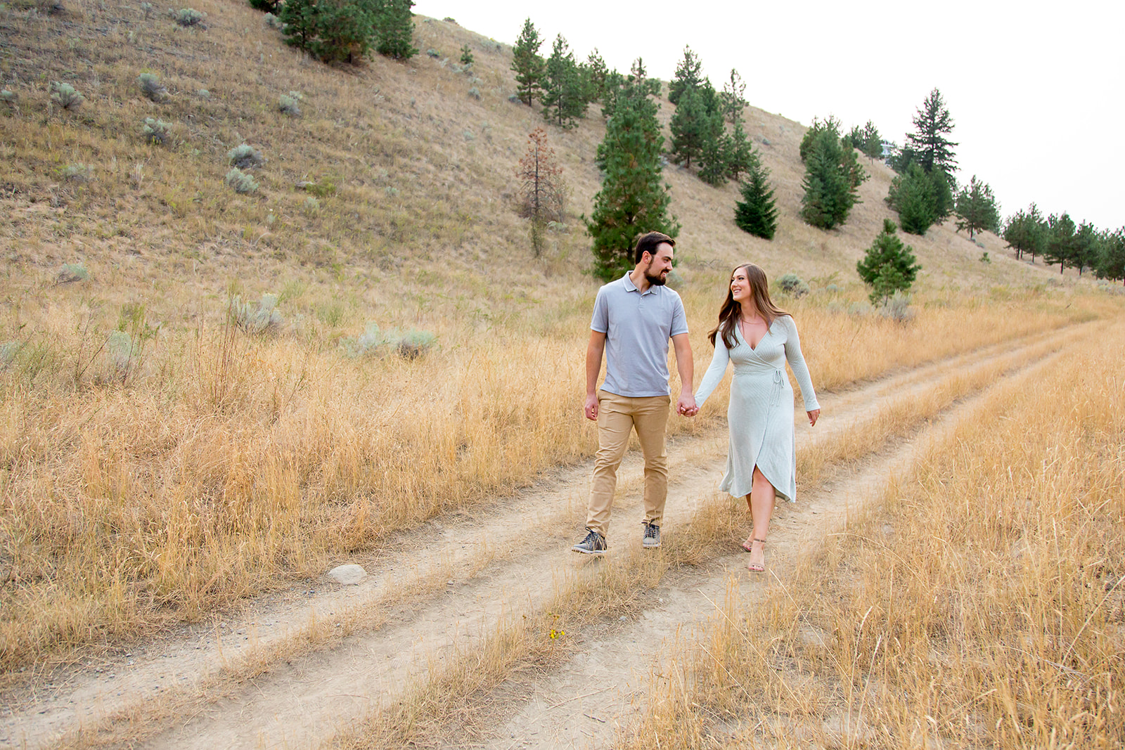 Young couple holds hands walking down a grassy path during their outdoor engagement session in Kamloops