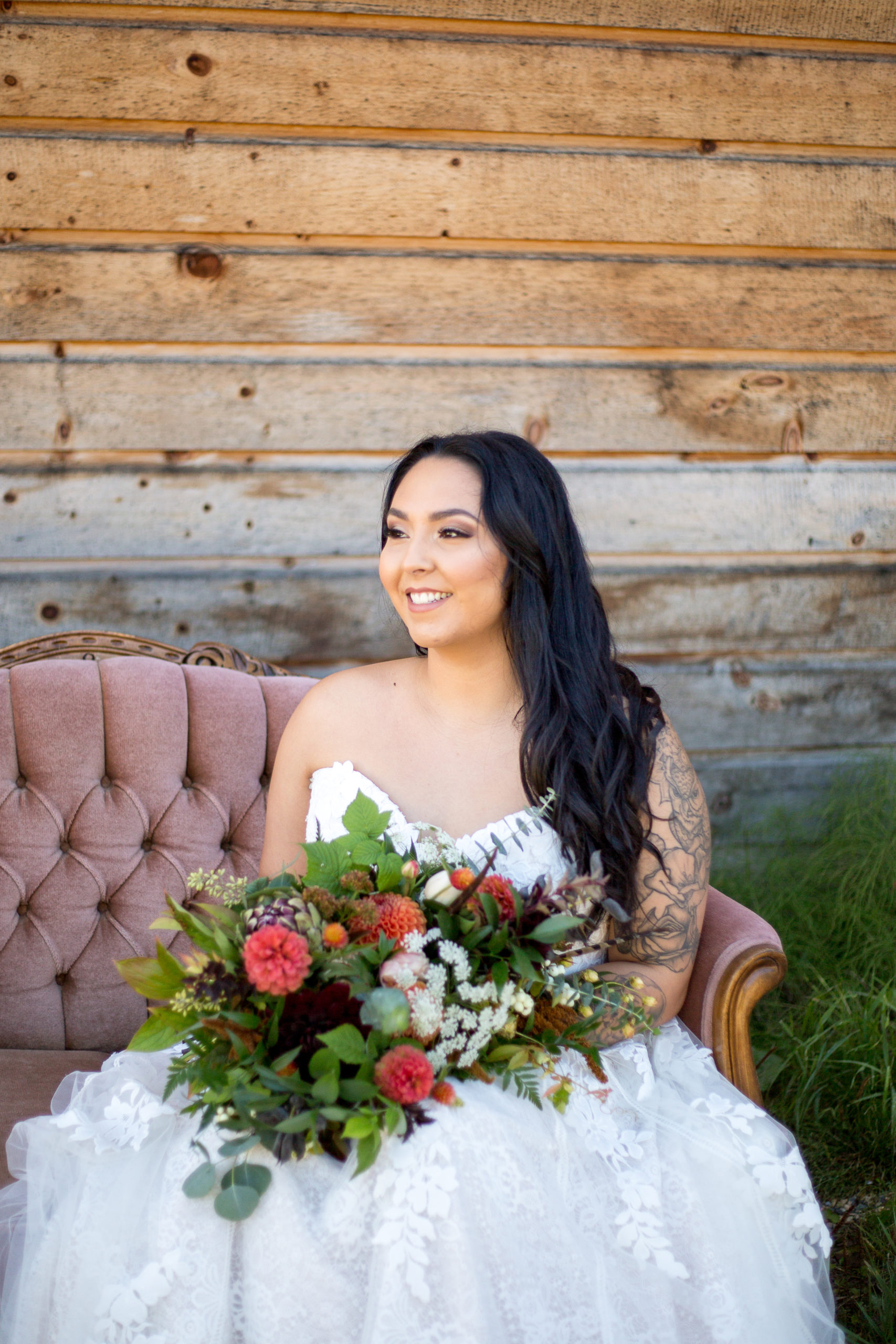 Bride sits on a blush velvet couch smiling holding her bouquet on her wedding day in BC