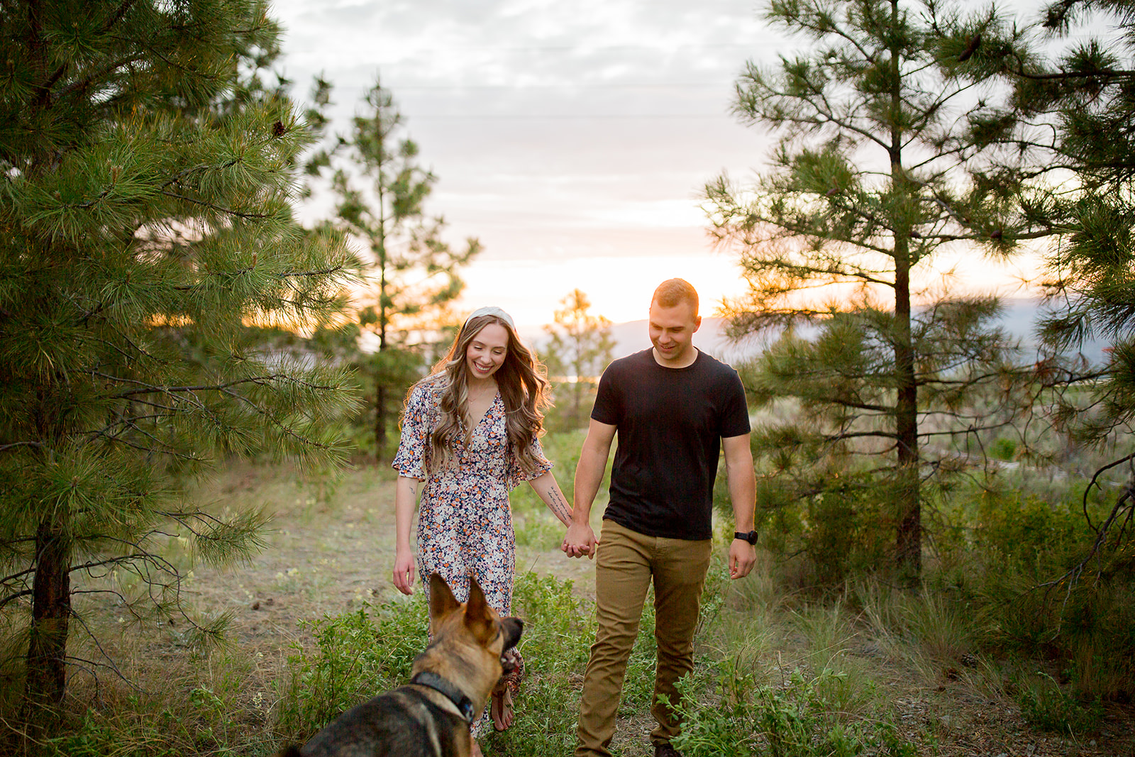 Newly engaged couple smiles warmly at their dog during their outdoor photoshoot in Kamloops at golden hour