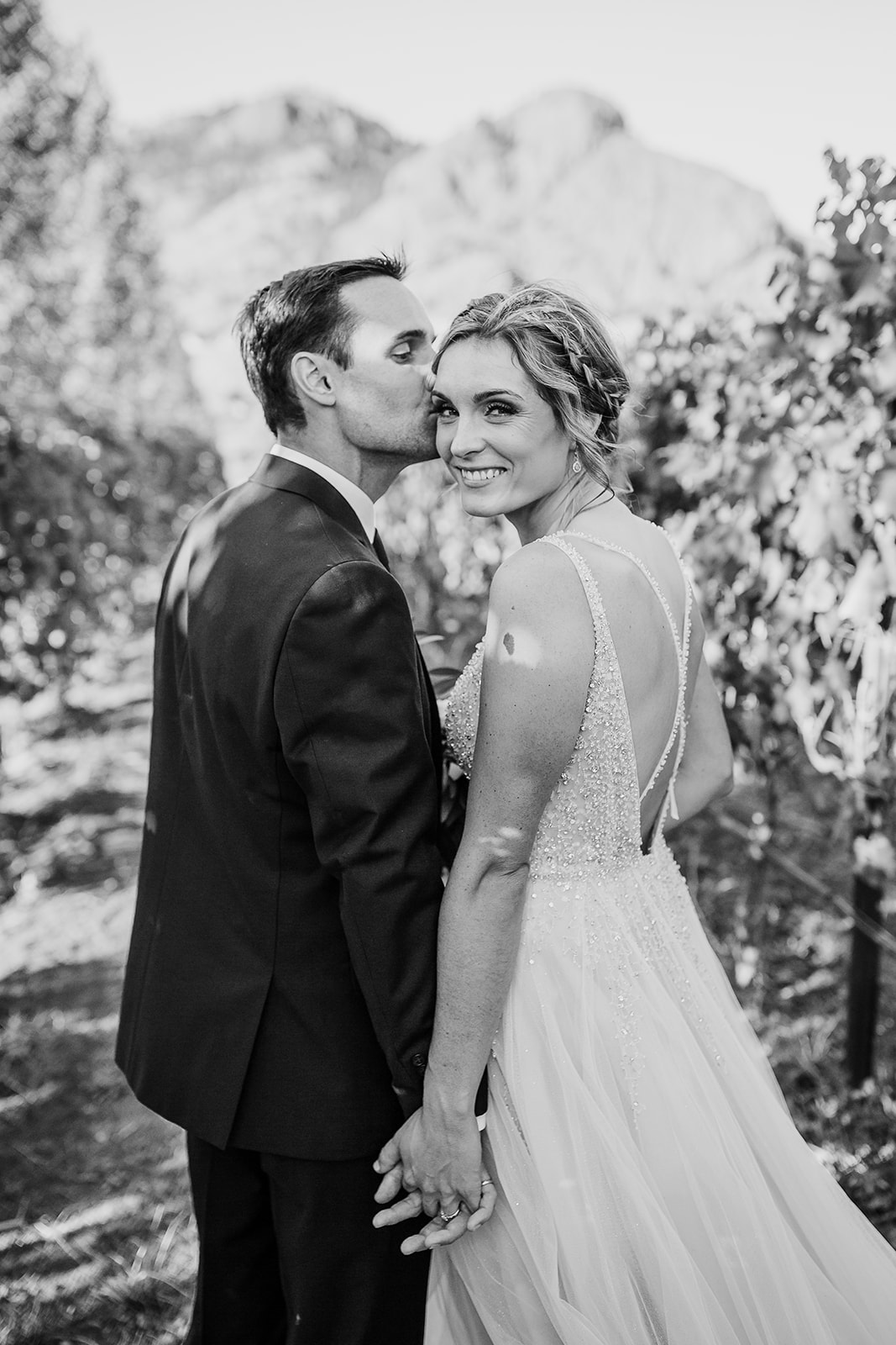 Black and white photo of a groom kissing his bride's cheek as they pose for their wedding photographer outside