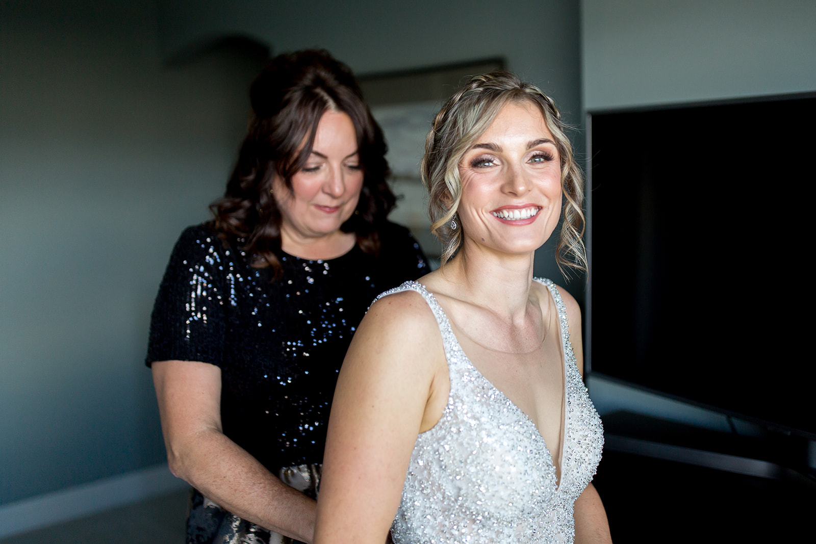 Bride smiles into the lens of the camera as her wedding photographer captures her being zipped into her dress on her wedding day