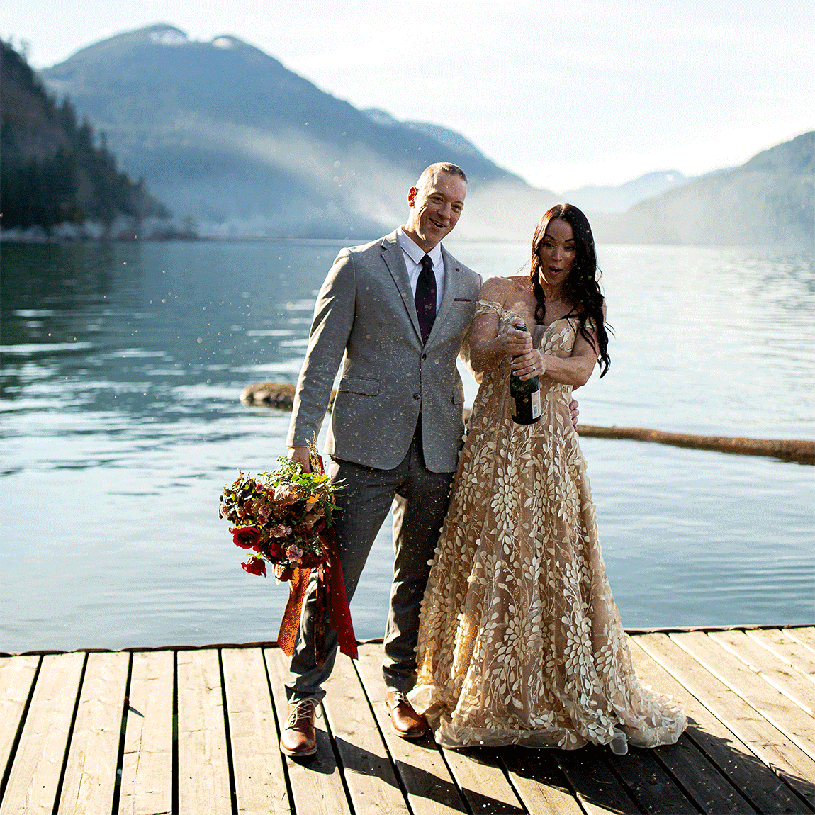 Gorgeous brunette in a gold dress sprays champagne next to a lake as her husband stands beside her smiling