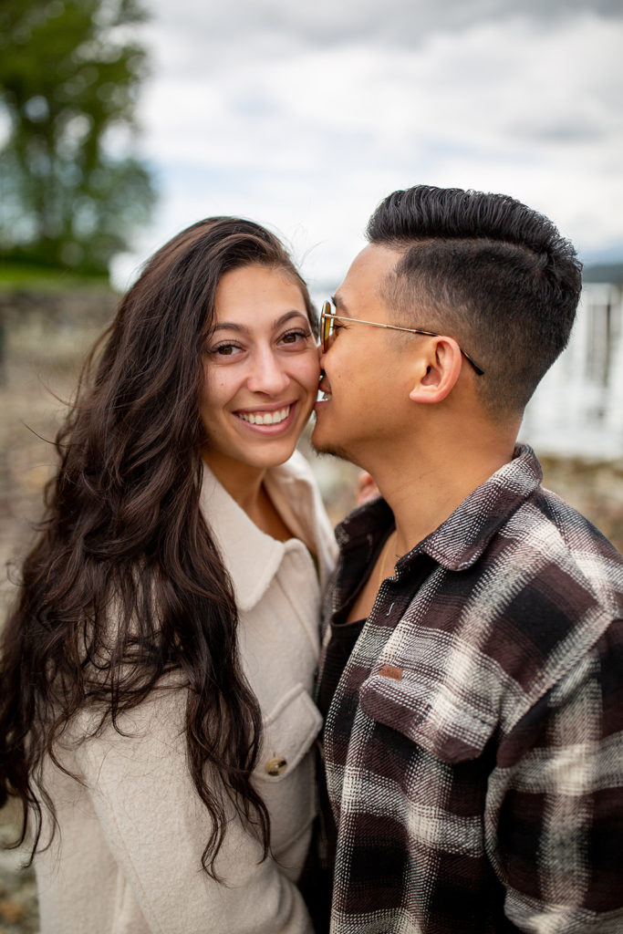 Man in plaid button-up shirt kisses the cheek of his partner as she smiles at the camera during their engagement photoshoot